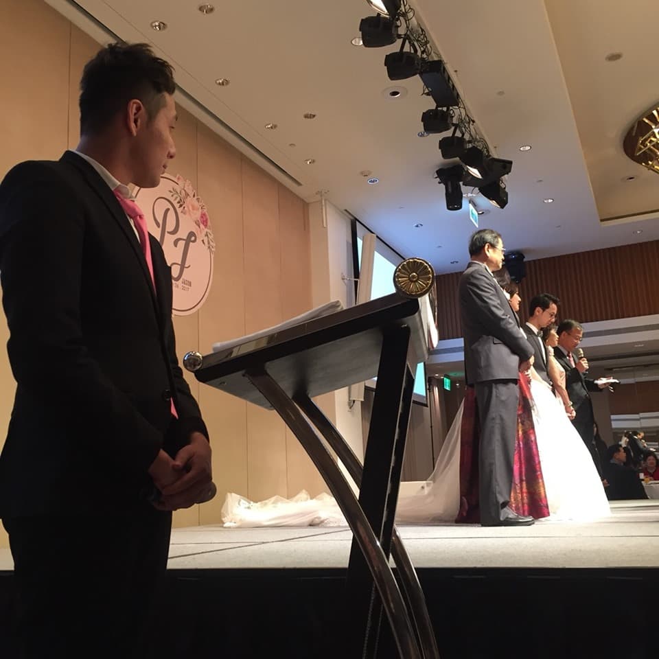 這張照片是一對新人舉行婚禮的場景,其中最重要的是站在講臺旁的男主角,他穿著黑色的西裝,打著領帶,看起來非常帥氣。