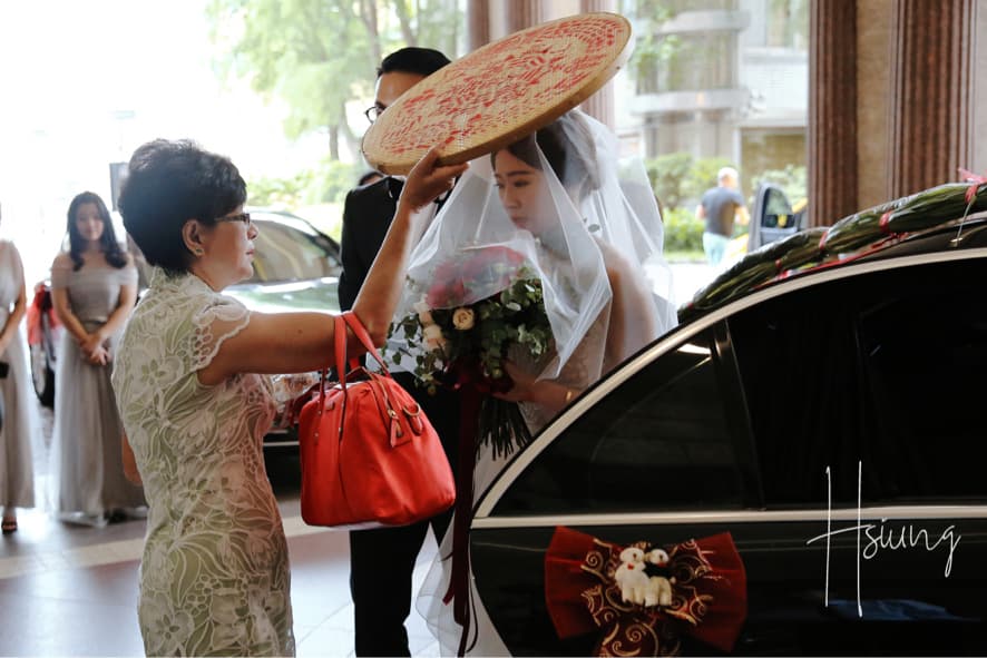 這張照片是一對新人準備上婚車的畫面。新郎站在車旁,新娘正要上車,手上拿著鮮花。一旁的新娘媽媽正為他們遮擋著。