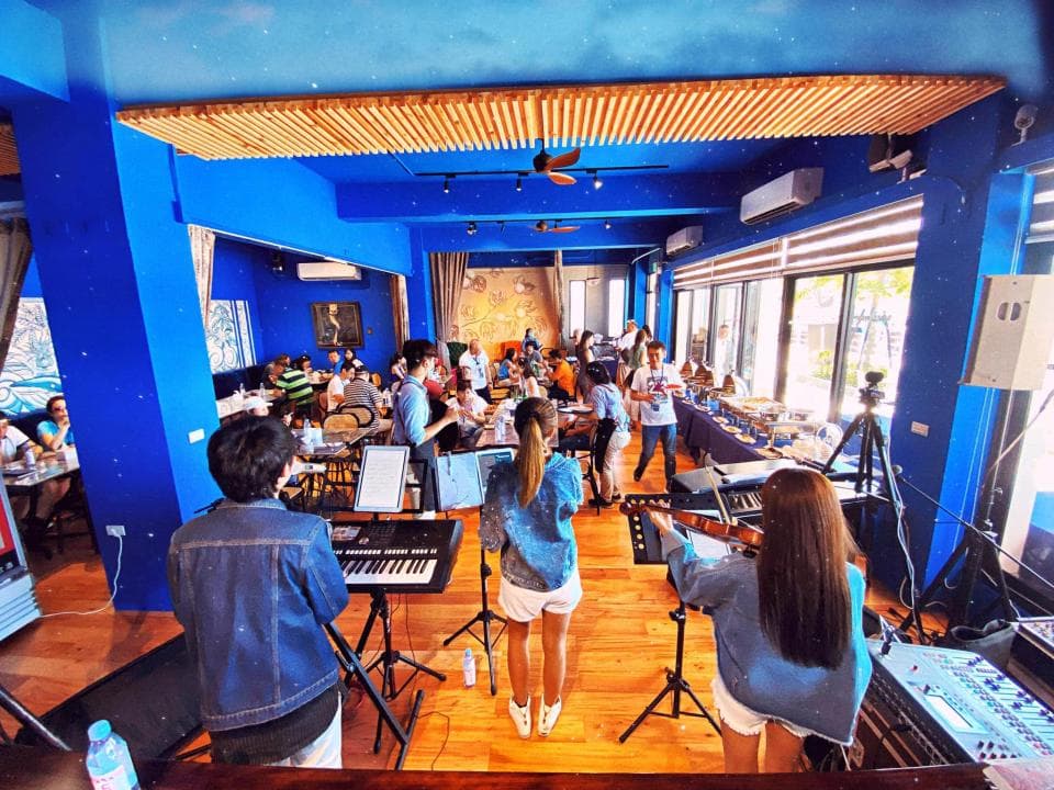 這張照片是一間以藍色為主的餐廳,餐廳裡有一群人正在演奏音樂。 舞臺上有一臺電子琴,兩位女生正演奏著不同的樂器,一位女生彈電子琴,另一位則演奏小提琴。