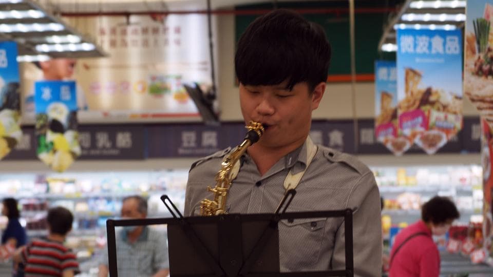 這張照片是一個年輕人在超市演奏薩克斯風。他專注地吹著樂器,看起來很投入。他身穿灰色襯衫,旁邊放著音樂譜架,背景是超市的貨架和商品。