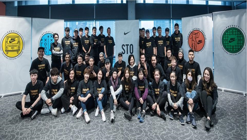 這張照片是一群人站在一面牆壁前,他們都穿著黑色T恤,上面印著金色字體。牆壁上有三個 Nike 標誌,以及一些文字。